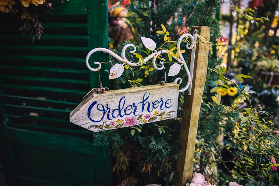 A rustic wooden garden sign with the words 'Order here' painted in blue cursive, hanging from a white metal scrollwork bracket. The sign is surrounded by lush green foliage and flowering plants, inclu