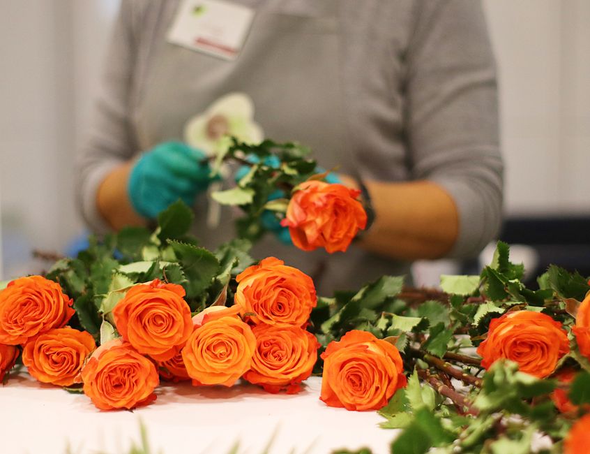 A florist in a grey long-sleeve top and blue gloves arranges a floral bouquet featuring vibrant orange roses with full, layered petals and lush green leaves on a white surface. Multiple stems of roses