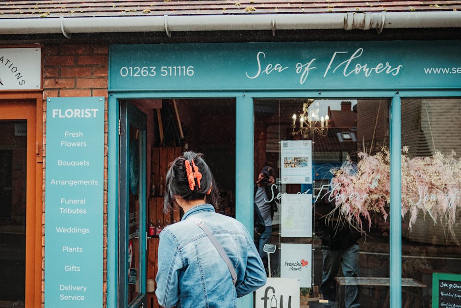A woman with short dark hair, secured with a large red clip, stands outside the floral shop 'Sea of Flowers' on Upper Street Barnsbury. She is wearing a light blue denim jacket and carrying a black sh