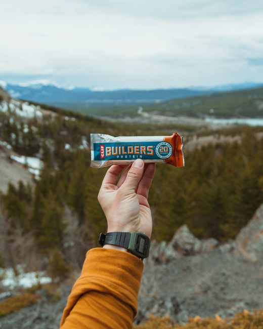 A person's hand holding a Clif Builder's Protein Bar in front of a scenic outdoor landscape. The hand, wearing a black wristwatch and an orange jacket sleeve, is positioned centrally. The protein bar 