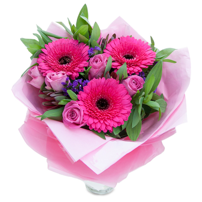 This composition captures the playful elegance of pink in a way that feels intimate and crafted by hand. Three vivacious fuchsia Gerbera daisies command the centre, their radiant petals forming concentric halos around deep, dark centres that give the bouquet a bold visual pulse. Nestled among them, roses in a softened lavender-pink or dusty-rose tone hold their petals tightly curled, the texture of each bloom like fine velvet waiting to unfurl - a sign of freshness and attentive timing. Scattered pockets of purple statice add dotted accents of cooler colour and a papery texture that contrasts with the roses' plushness. A generous framework of greenery - broad salal-like leaves and the rounded, aromatic stems of eucalyptus - supports the blooms and lends a garden-fresh scent that offsets the roses' gentle perfume. The bouquet is wrapped with care: a pale pink matte tissue creates a tender inner layer, while a sheer, slightly crinkled fuchsia-pink cellophane overlays it, catching the light with a soft shimmer and reinforcing the overall palette. The wrapping is folded artfully around the stems, and the top rim of a clear glass vase is visible at the base, suggesting this arrangement is prepared for immediate placement on a mantel or as a focal table accent. Evoking the quiet charm of Barnsbury Square or a sunny morning stroll near Upper Street, this bouquet is a warm, thoughtful choice for birthdays, anniversaries, or a simple, uplifting 'just because' gift.